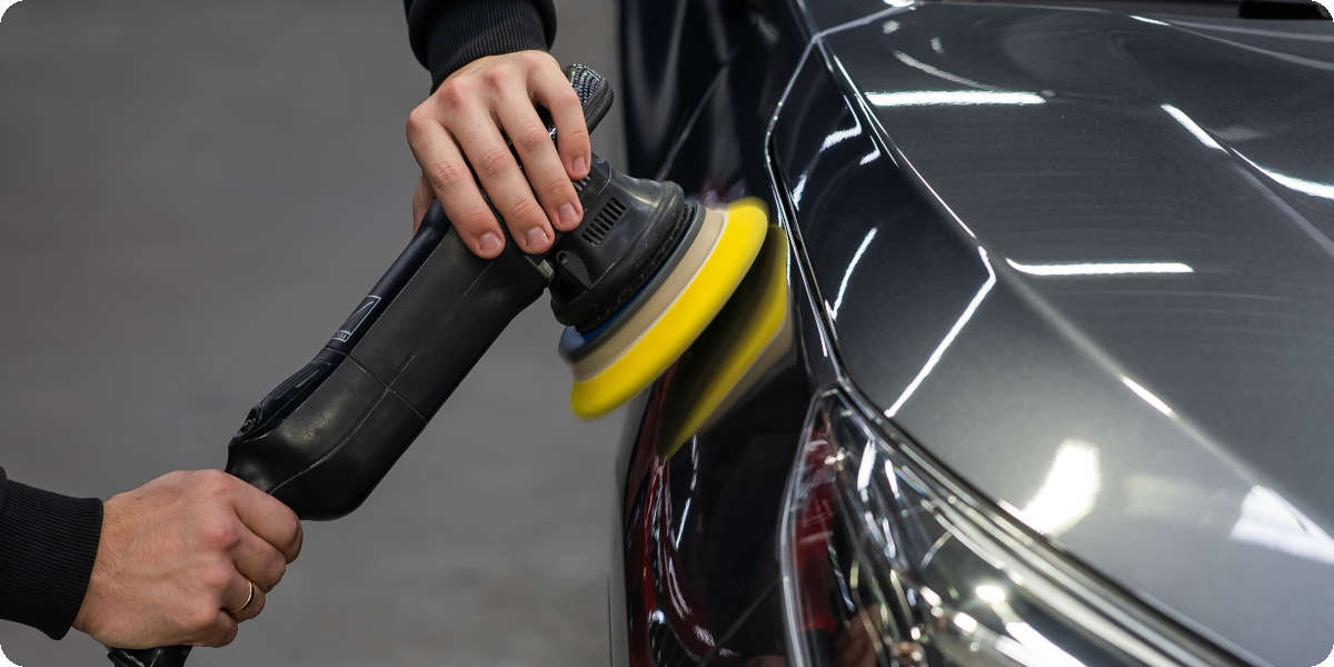 Nahaufnahme von zwei Händen, die ein graues Auto mit einer Maschine polieren.