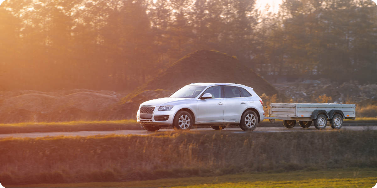 Ein silbernes Auto zieht einen Anh&auml;nger auf einer Landstra&szlig;e im Sonnenuntergang.